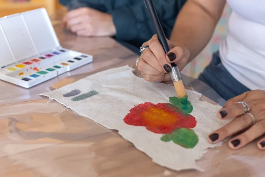 Gros plan d'une main en train de peindre à l'aquarelle sur un tissu en coton lors d'un essai lors d'un atelier d'initiation aquarelle et broderie