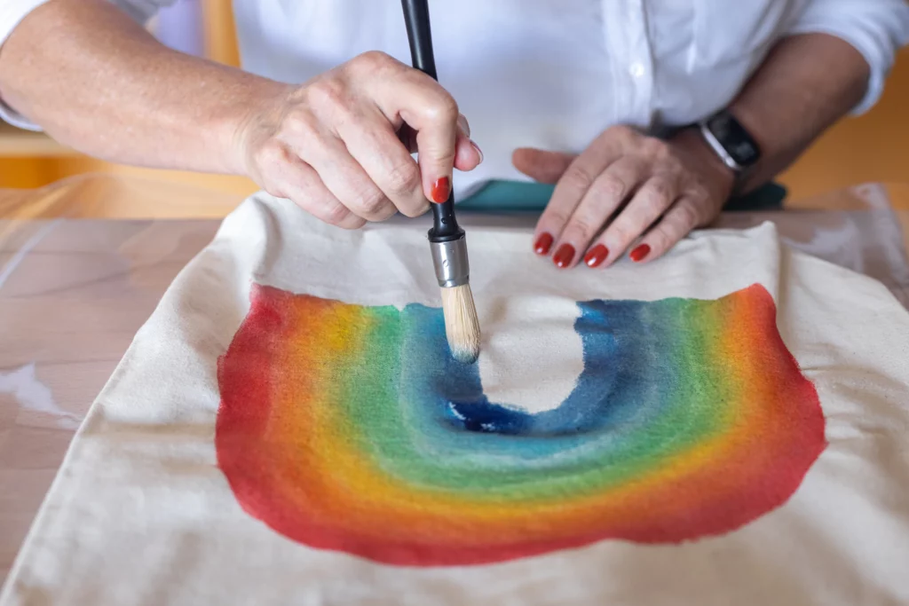 Main peignant à l'aquarelle sur un tissu de tote bag, représentant un arc-en-ciel fait lors d'un atelier d'initiation