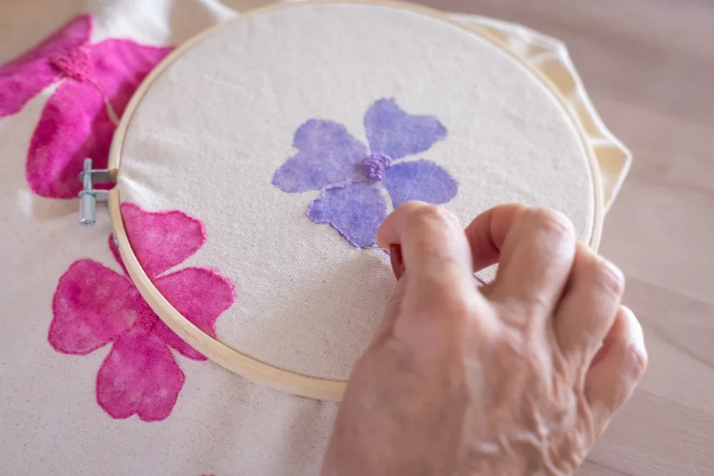 Gros plan d'une main en train de faire de la broderie main autour d'une fleur faite à l'aquarelle sur du tissu lors d'un atelier créatif