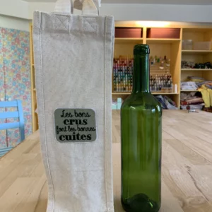 Porte bouteille posé à côté d'une bouteille de vin vide, avec une broderie "les bons crus font les bonnes cuites"