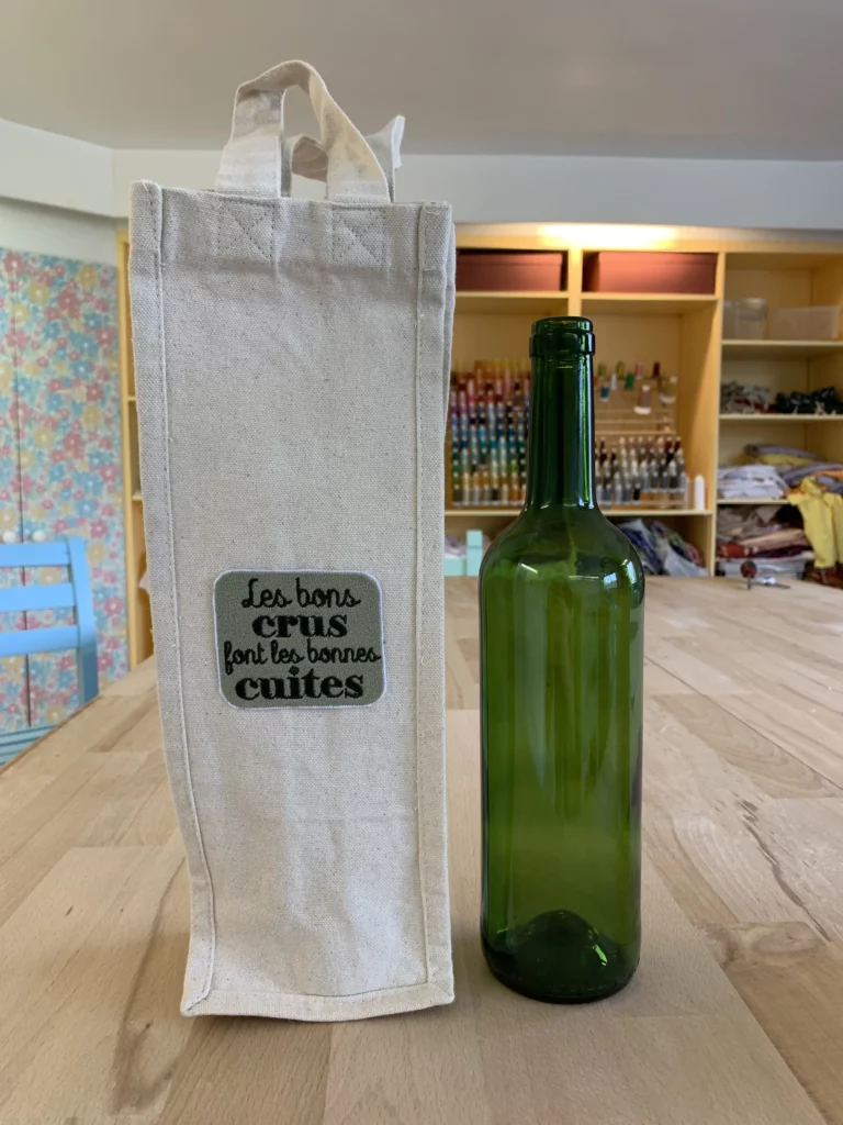 Porte bouteille posé à côté d'une bouteille de vin vide, avec une broderie "les bons crus font les bonnes cuites"