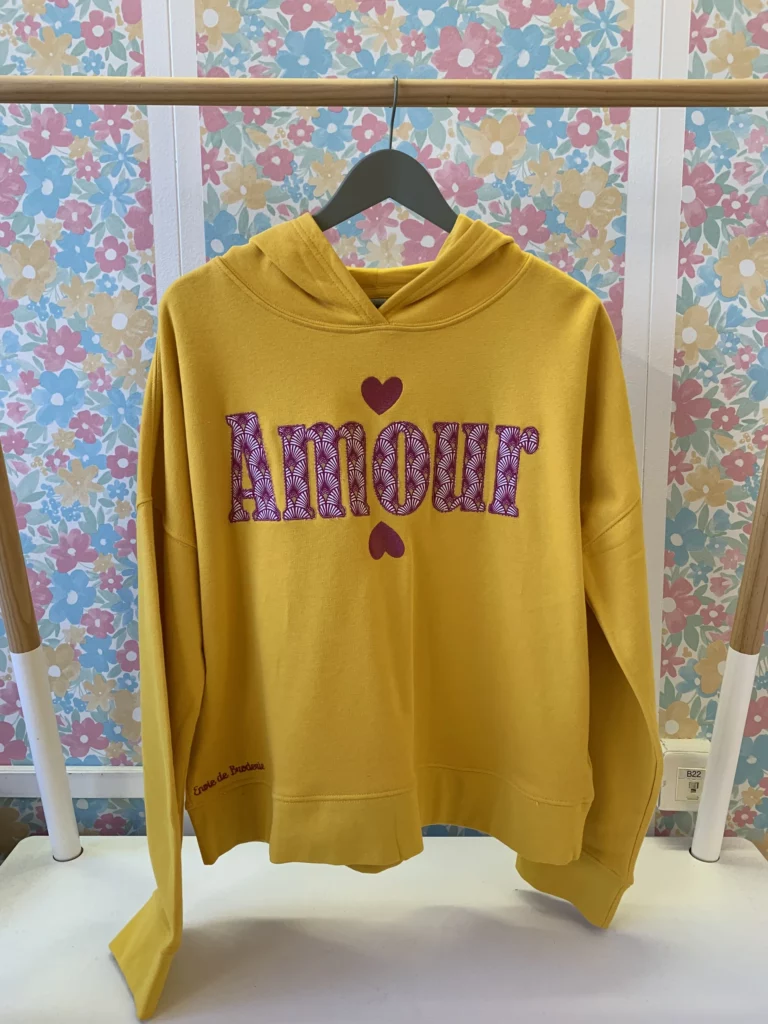 Vue de face du sweat jaune en pièce unique, brodé "Amour" avec deux coeurs brodés, dont l'intérieur des lettres contient un tissu rouge