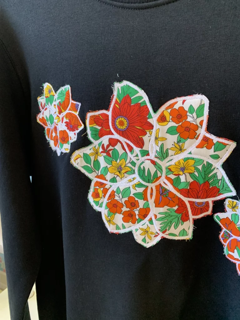 Gros plan de trois fleurs brodés sur un sweat-shirt noir, donc l'intérieur des fleurs contient un tissu dans les tons rouges et blanc avec pleins de fleurs.