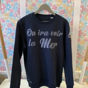 Vue de face du sweat-shirt en pièce unique brodé "on ira voir la mer" dont l'intérieur des lettres contient un tissu en marinière