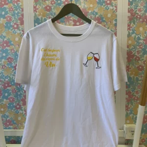 T-shirt blanc vue de face avec une broderie de deux verres de vin côté coeur, ainsi qu'un texte "c'est toujours l'heure du verre de vin" en contre-coeur