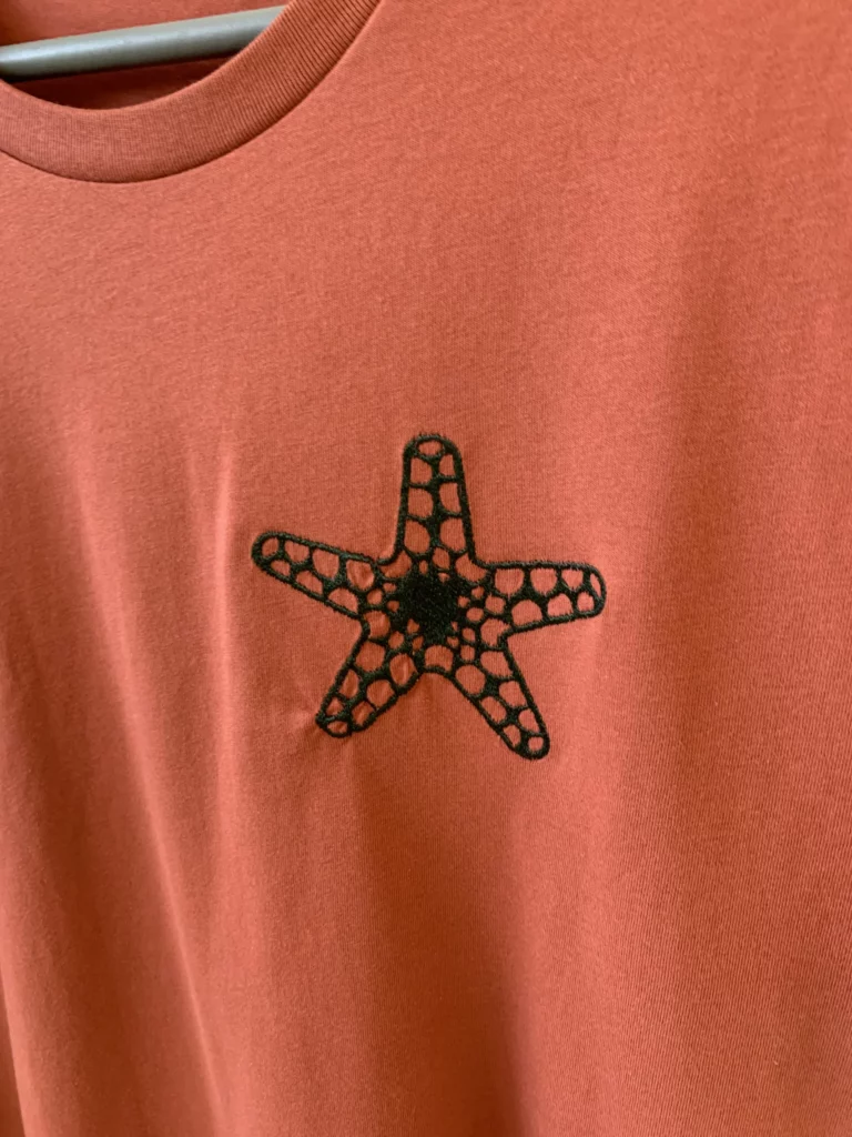 Gros plan d'une broderie en forme d'étoiles de mer, de couleur verre forêt, sur un t-shirt de couleur orange