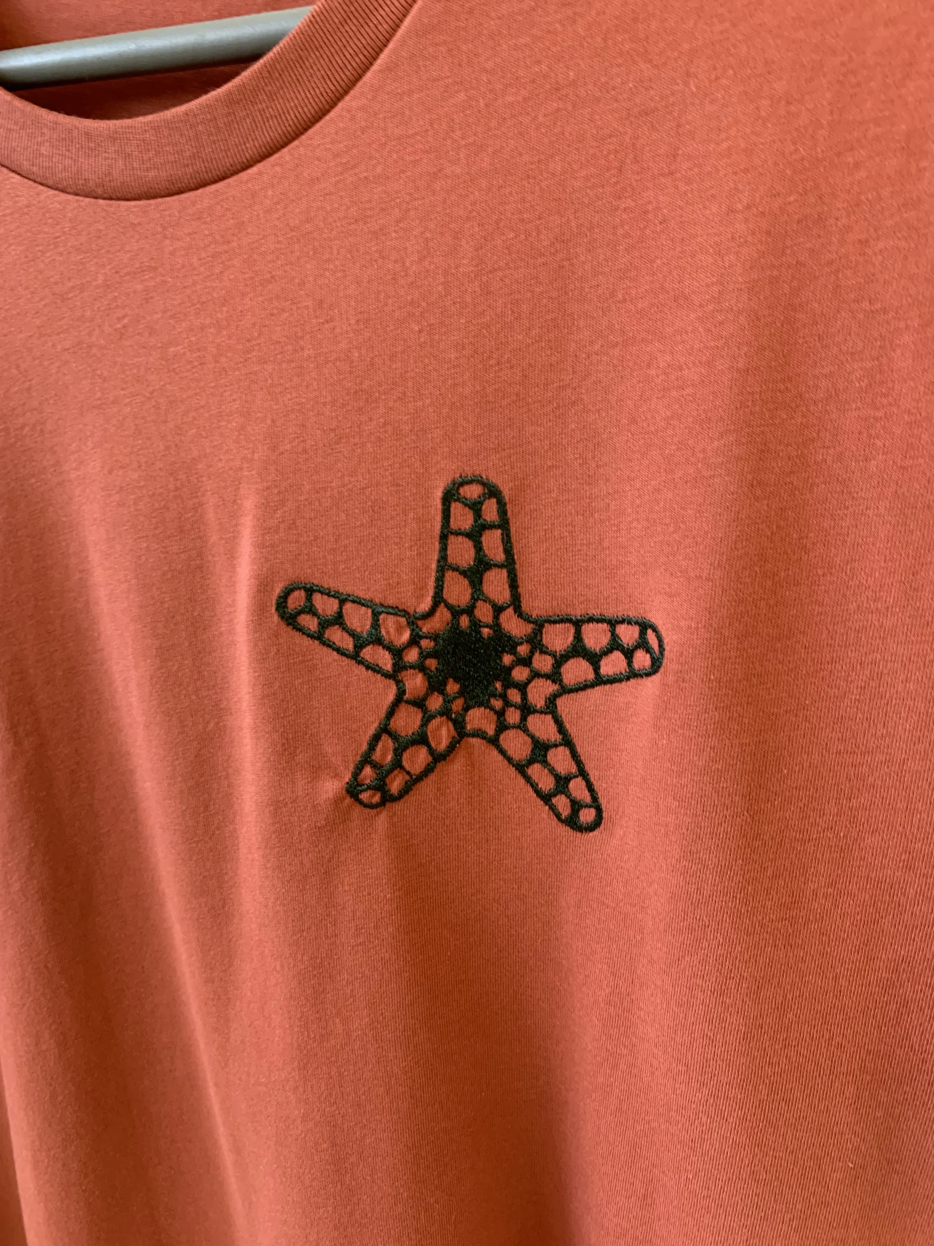 Gros plan d'une broderie en forme d'étoiles de mer, de couleur verre forêt, sur un t-shirt de couleur orange