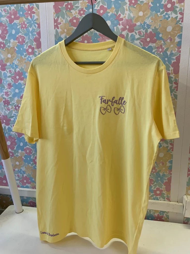 T-shirt jaune en coton biologique avec une broderie violette représentant deux pâtes italiennes, ainsi que le mot "farfalle"