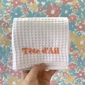 Broderie du mot "tête d'ail" en orange sur un torchon de cuisine blanc